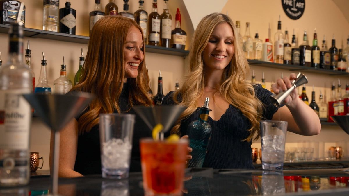 Two women behind a bar preparing cocktails with bottles and glasses in the background at Fine Art Bartending School