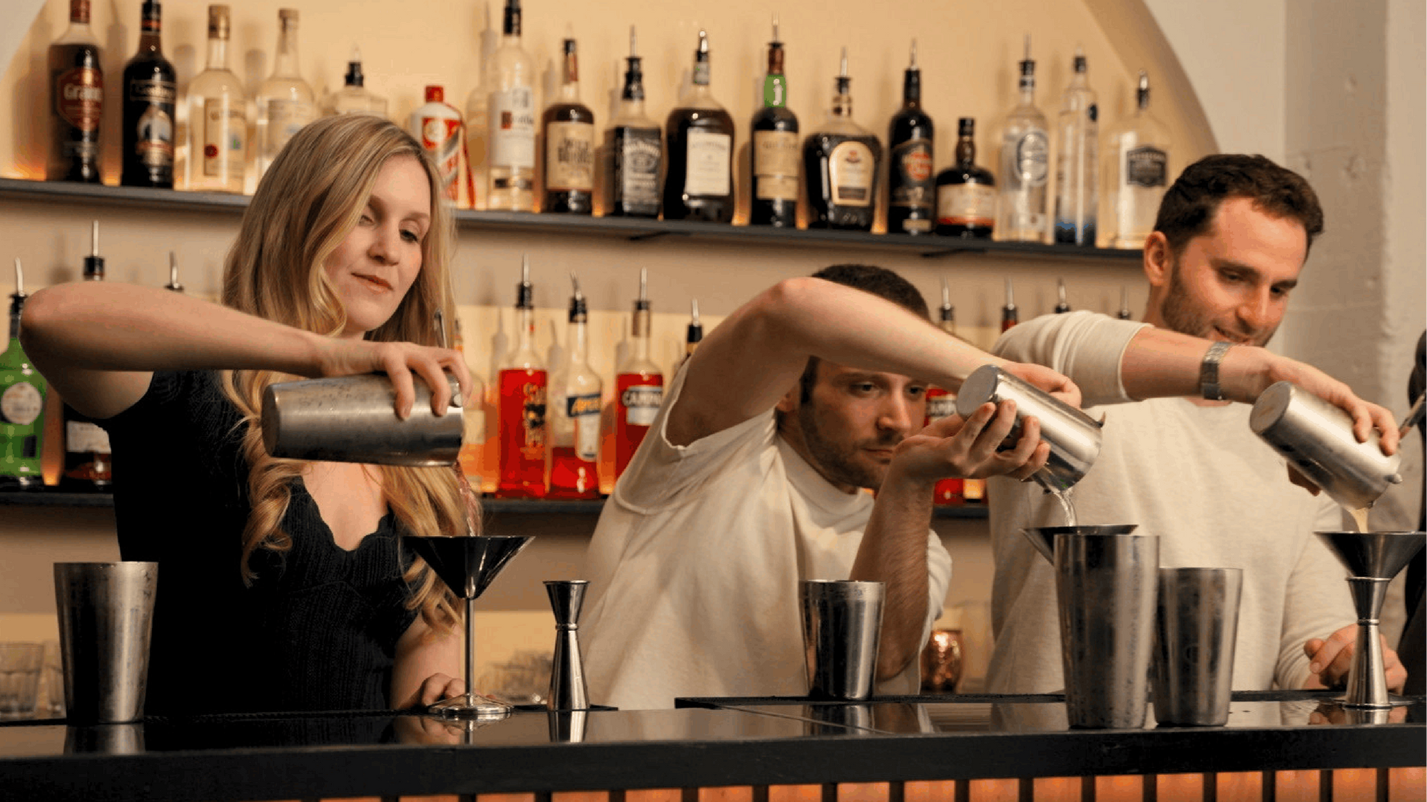 Three baristas pour drinks behind a bar counter, with shelves of liquor bottles in the background.