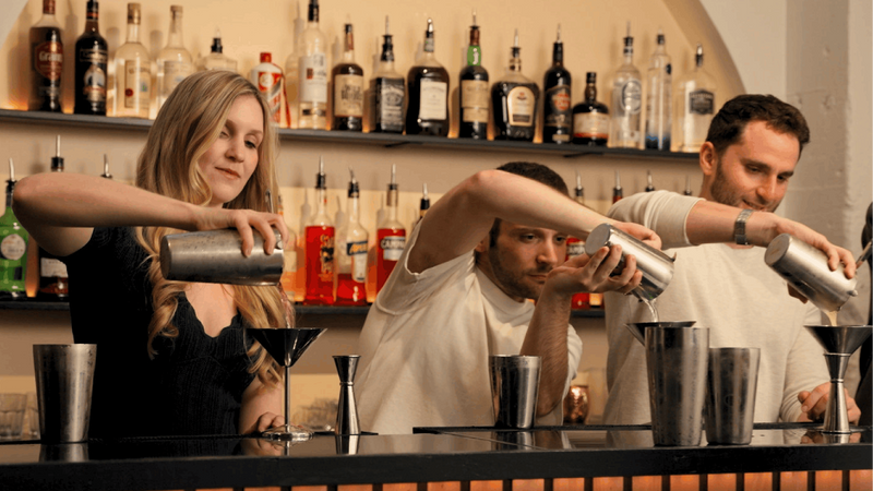 Three baristas pour drinks behind a bar counter, with shelves of liquor bottles in the background.