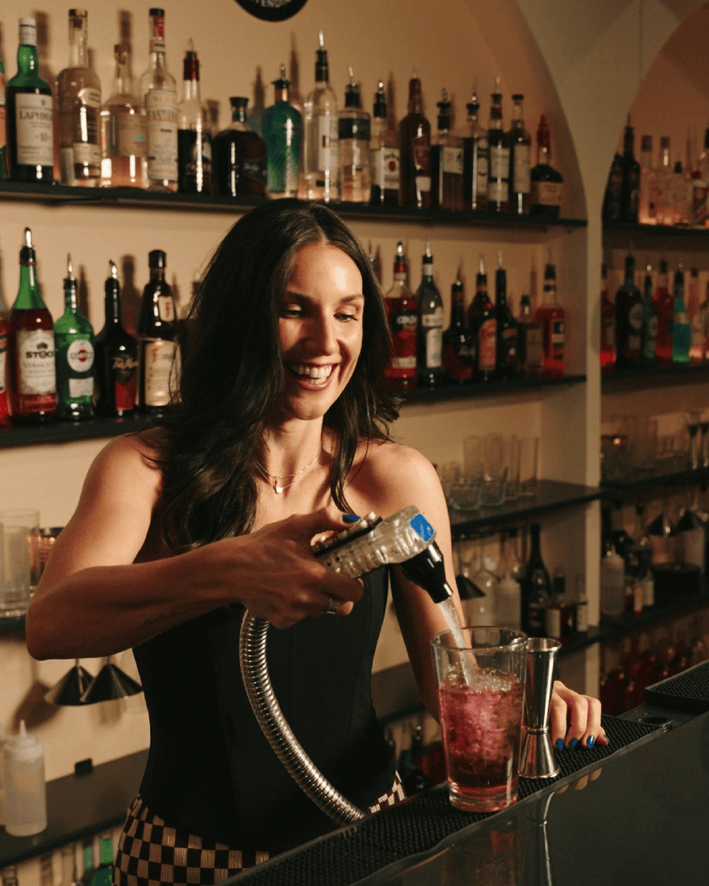 A smiling woman bartender in a black top and checkered skirt uses a metal muddler tool to press down a pink cocktail in a glass on a bar counter, with shelves of liquor bottles in the background.